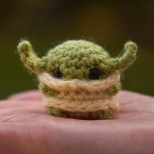 May include: A small green crocheted creature with large ears and a cream-coloured scarf. The creature has black eyes and a friendly expression.