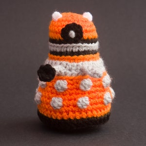 May include: A crocheted orange and grey robot toy with black details. The robot has a round body with a white circle for an eye and a black circle in the centre.