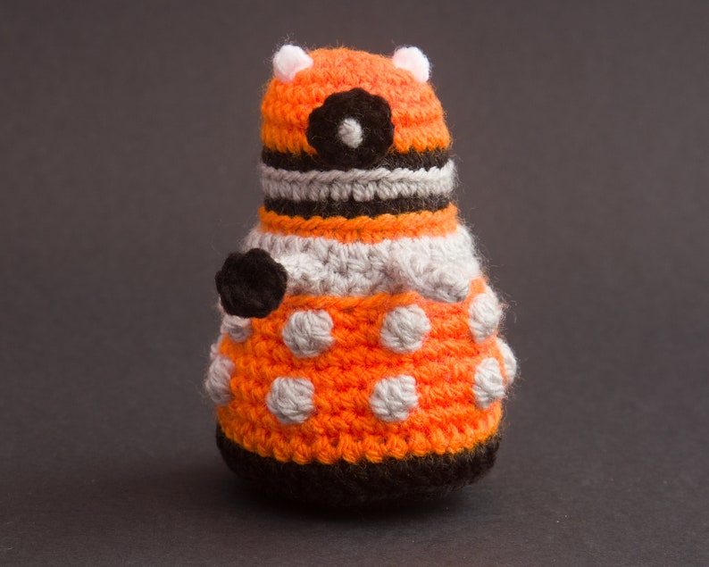May include: A crocheted orange and grey robot toy with black details. The robot has a round body with a white circle for an eye and a black circle in the centre.