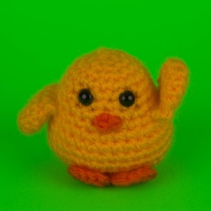 May include: A yellow crocheted chick toy with black eyes and an orange beak. The chick is standing on a green background.