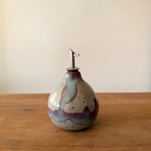 May include: A ceramic oil dispenser with a bulbous shape and a stainless steel spout. The stoneware bottle features a speckled, glazed finish with shades of gray, teal, and burgundy. The dispenser is designed for easy pouring and adds a touch of rustic elegance.