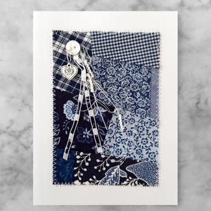 May include: A handmade card with a patchwork design featuring various shades of blue fabric. The card has a white button and a silver heart charm.