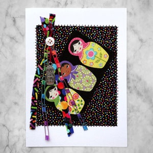 May include: A white card with a black fabric background with colorful polka dots.  There are three colorful Russian nesting dolls with faces and floral patterns on the fabric.  The fabric is trimmed with black thread and has colorful frayed ribbons hanging from the bottom.