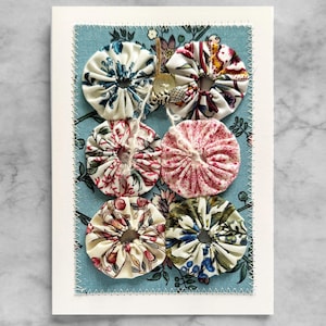 May include: A handmade greeting card featuring six fabric yo-yos in various floral patterns. The yo-yos are attached to a light blue fabric with a floral design, which is stitched onto a white card. A small charm and button are attached.