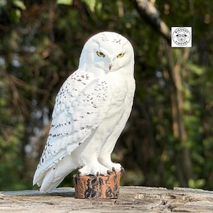 May include: A white snowy owl figurine with black speckled markings on its wings and back, featuring bright yellow eyes and a sharp beak. The owl is perched on a small, textured brown cylindrical base. A logo with the text BIGSHOT TINKERS is visible.
