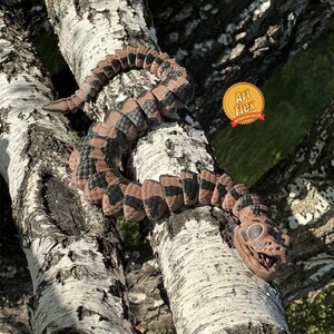 May include: A segmented, articulated snake figure with a brown and black patterned body and a detailed skull-like head. The snake is coiled along a textured tree branch. A circular orange and yellow badge with the text Art Flex is visible.