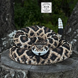 Puede incluir: Una figura de serpiente de cascabel enroscada con un patrón detallado de escamas negras, bronceadas y blancas, con un cascabel levantado en la cola y una lengua que sobresale. Un logotipo cuadrado blanco con el texto BIGSHOT TINKERS es visible.