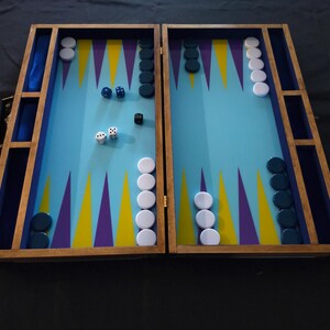 Peut inclure: Un jeu de backgammon en bois ouvert, avec une surface de jeu bleu clair ornée de pointes triangulaires violettes et jaunes. Le jeu affiche des pions blancs et bleu foncé, des dés et un cube de doublement noir. Le cadre en bois a une poignée.