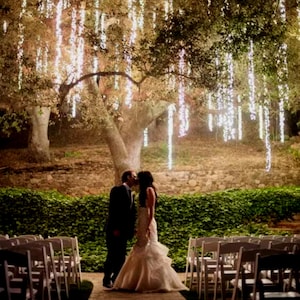 May include: A wedding ceremony taking place outdoors under a tree draped with white string lights. The bride and groom are kissing in front of a row of white chairs.