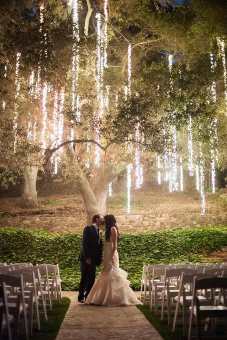 Pu&ograve; includere: Una cerimonia di matrimonio all'aperto sotto un albero con luci bianche a catena appese ai rami. La sposa e lo sposo si baciano al centro della navata, con sedie bianche allineate su entrambi i lati.