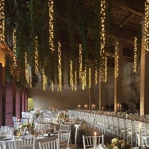 May include: A large, rustic venue decorated with hanging greenery and string lights for a wedding reception. White chairs and tables are set up for guests, with candles and floral centerpieces.