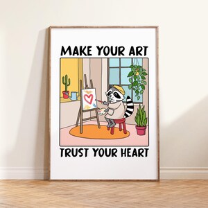 May include: A framed art print featuring a cartoon raccoon artist painting a heart on a canvas. The print includes the text "MAKE YOUR ART" above and "TRUST YOUR HEART" below the image. The raccoon is wearing a beret and a painter's smock.