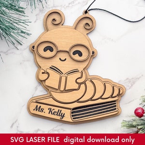 Puede incluir: Adorno de madera con un gusano de dibujos animados con gafas leyendo un libro. El gusano está posado sobre una pila de libros con el nombre "Ms. Kelly" grabado. El adorno tiene una cuerda para colgar.