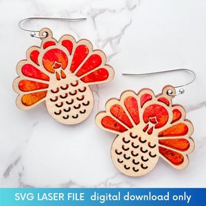 May include: A pair of wooden turkey earrings with red, orange, and yellow accents. The earrings have a laser-cut design with a natural wood finish and silver-colored ear wires. The text "SVG LASER FILE digital download only" is at the bottom.