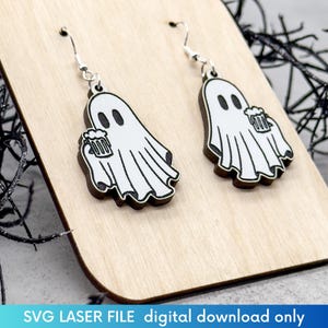 May include: A pair of wooden earrings featuring a ghost holding a beer. The earrings are white with black outlines and are hanging from silver hooks.
