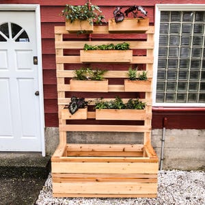 May include: A wooden vertical garden planter with multiple shelves and a large planter box at the bottom. The planter is made of natural wood and has a rustic look. It is filled with various plants and flowers.