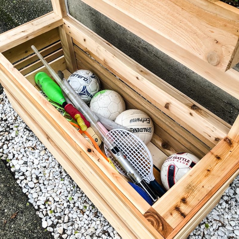 May include: A wooden storage box with a hinged lid, filled with sports equipment, including balls, bats, and a tennis racket. The box is made of natural wood and has a rustic look.