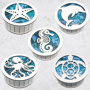 Pode incluir: Cinco caixas redondas de madeira com fundo branco e design de ondas do mar azul e branco. Cada caixa apresenta um recorte de criatura marinha diferente: uma estrela do mar, um cavalo-marinho, um golfinho, um polvo e uma tartaruga marinha.