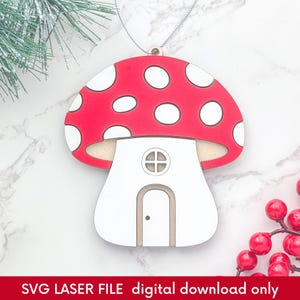 Op de afbeelding: Rode en witte paddenstoelvormige kerstversiering met een wit huisontwerp en een kleine deur. De versiering is gemaakt van hout en heeft een touwtje om op te hangen.