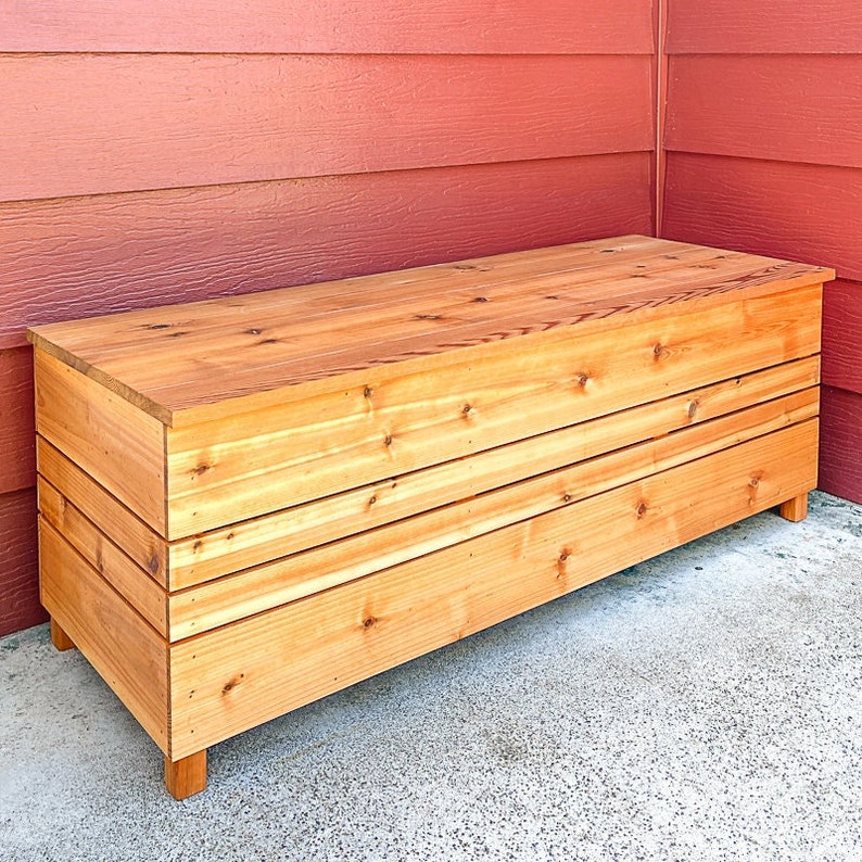 May include: A wooden storage bench with a natural wood finish. The bench has a hinged lid and is supported by four legs.