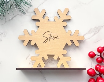 May include: A wooden snowflake-shaped place card holder with the name "Steve" engraved in a script font. The snowflake is light brown and sits on a dark brown base. Decorative greenery and red berries are in the background.