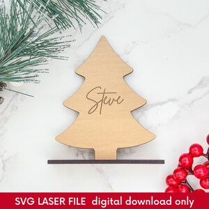 Puede incluir: Una tarjeta de lugar de madera con forma de árbol de Navidad con el nombre "Steve" grabado. El árbol es de color marrón claro y se asienta sobre una base marrón oscuro. La imagen incluye una ramita de pino y bayas rojas.