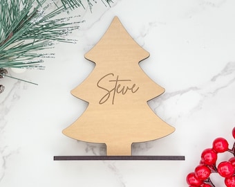 May include: A wooden Christmas tree-shaped place card with the name "Steve" engraved on it. The tree is light brown and sits on a dark brown base. The image includes a sprig of pine and red berries.