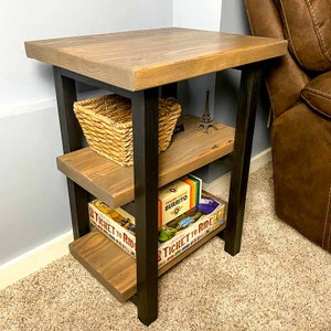 Puede incluir: Una mesa auxiliar de madera de tres niveles con patas de metal negro. La repisa superior tiene una cesta tejida y una pequeña figura de la Torre Eiffel. La repisa del medio tiene un juego de mesa llamado "The One-Eyed Jack Burrito". La repisa inferior tiene un juego de mesa llamado "Ticket to Ride".