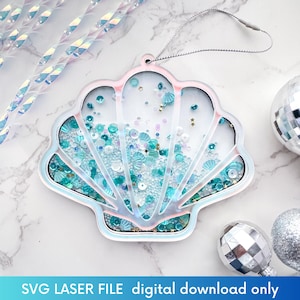 May include: A white iridescent seashell ornament with a blue and white glitter design. The ornament has a string loop for hanging. SVG LASER FILE digital download only.