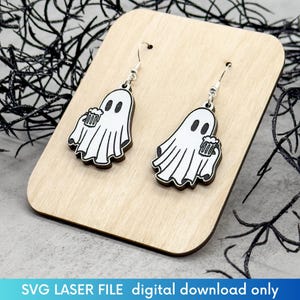 May include: A pair of wooden earrings featuring a ghost holding a beer mug. The earrings are white with black outlines and are hanging from silver hooks.