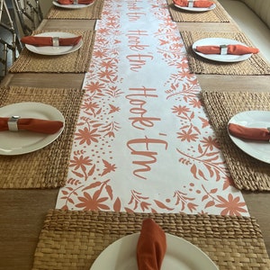 May include: A dining table set for a meal, featuring a white table runner with the text "Hook 'Em" in orange. The runner is decorated with floral patterns. Each place setting includes a white plate, orange napkin, and silver napkin ring.