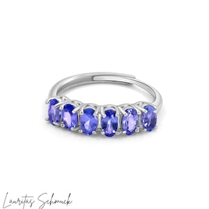 Könnte beinhalten: Silberfarbener Ring mit sechs ovalen, blauen Edelsteinen. Der Ring ist verstellbar. Die Edelsteine sind in einer Reihe angeordnet. Der Text "Lauritas Schmuck" steht in Kursivschrift unten. Weißer Hintergrund.