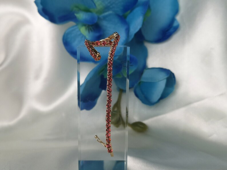 May include: A gold hair clip with a red rhinestone design. The clip is shaped like a curved line.