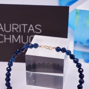 May include: A dark blue gemstone bracelet with a gold clasp. The bracelet is on a clear display stand. The text "LAURITAS SCHMUC" is visible in the background.