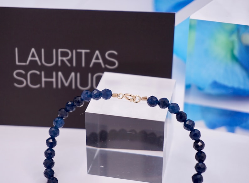 May include: A dark blue gemstone bracelet with a gold clasp. The bracelet is on a clear display stand. The text "LAURITAS SCHMUC" is visible in the background.