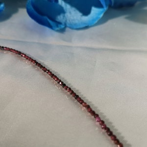 May include: A close-up of a delicate necklace made of small, dark red beads. The necklace is lying on a white fabric surface.
