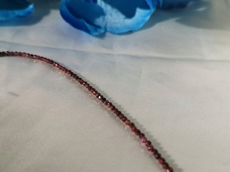 May include: A close-up of a delicate necklace made of small, dark red beads. The necklace is lying on a white fabric surface.