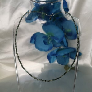 May include: A delicate necklace made of small, dark green beads. The necklace is displayed on a glass stand with blue flowers.