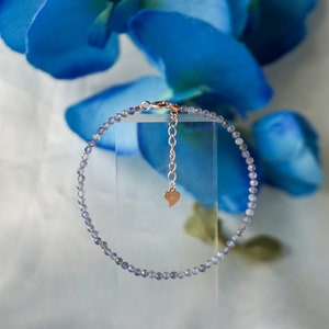 May include: A delicate bracelet made of small, light purple beads with a rose gold chain and heart charm.
