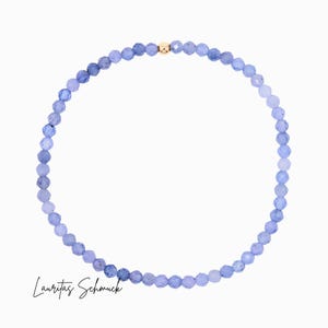 Può includere: Una delicata collana con piccole perline blu chiaro sfaccettate. Un'unica perla color oro è centrata sulla collana. La collana è esposta su sfondo bianco. Il testo "Lauritas Schmuck" è in basso a sinistra.