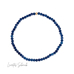 Könnte beinhalten: Ein zartes, dunkelblaues Perlenarmband mit einer goldfarbenen Akzentperle. Das Armband ist kreisförmig und liegt auf weißem Hintergrund. Der Text "Lauritas Schmuck" ist unten in Schreibschrift geschrieben.