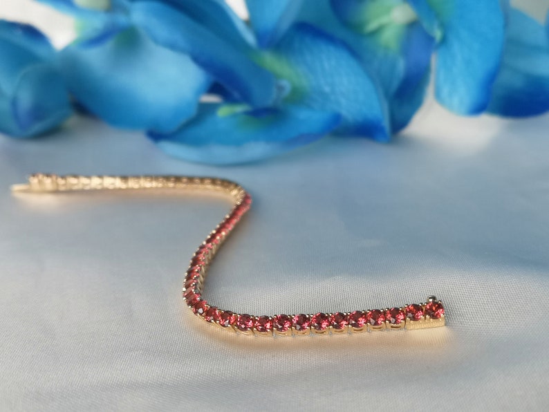 May include: A gold tennis bracelet with a row of small, round, red gemstones.