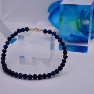 May include: A dark blue beaded bracelet with a gold clasp. The bracelet is made of small, round beads and is lying on a white surface.