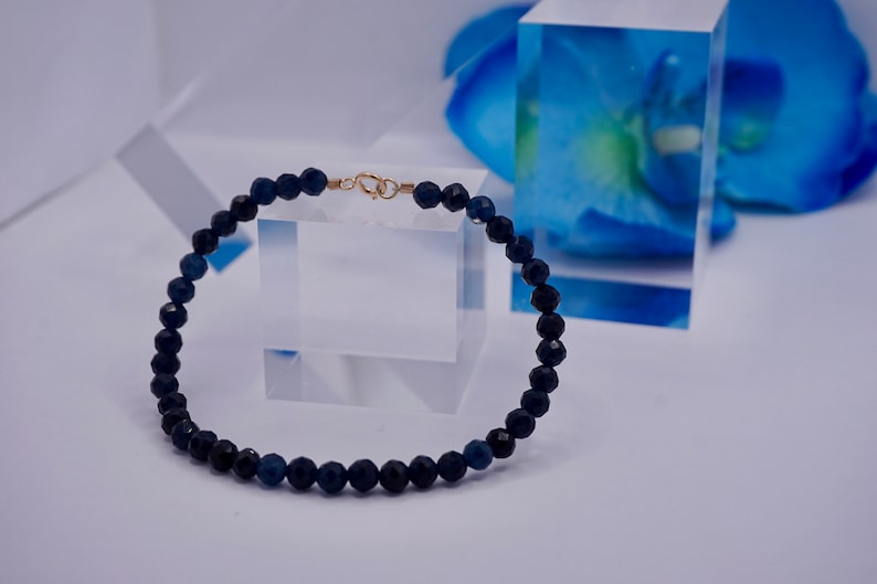 May include: A dark blue beaded bracelet with a gold clasp. The bracelet is made of small, round beads and is lying on a white surface.