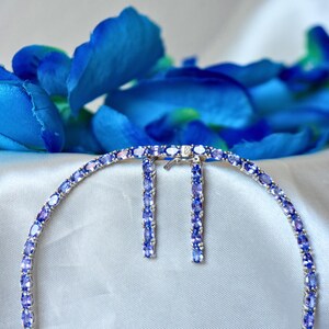 Lila Tansanit Edelstein in Tennishalskette und Ohrringe gefasst. Im Hintergrund eine Blaue Blume.