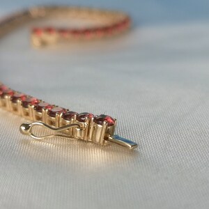 May include: A gold bracelet with a row of small, red gemstones. The bracelet has a clasp closure.