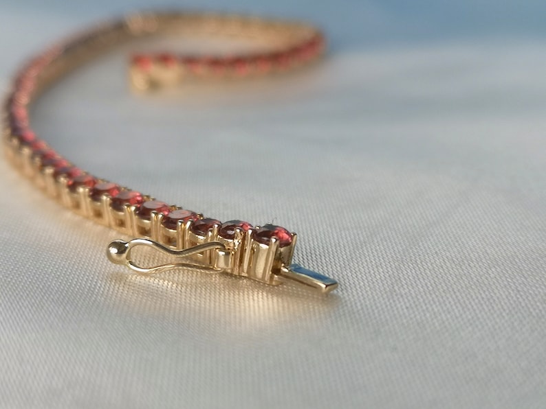 May include: A gold bracelet with a row of small, red gemstones. The bracelet has a clasp closure.