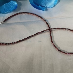 May include: A delicate necklace made of small, dark red garnet beads. The necklace is strung on a silver chain and has a small heart-shaped charm.