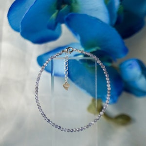 May include: A delicate silver chain bracelet with a heart charm and small, light purple beads.