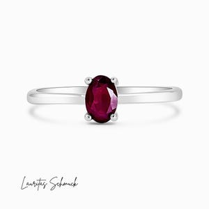 Può includere: Un anello d'argento con un rubino ovale di colore rosso intenso. La gemma è tenuta da quattro griffe. La fascia è sottile e lucida. Il testo "Lauritas Schmuck" è scritto in corsivo.